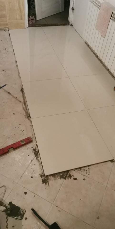 Ułożone jasne płytki podłogowe w trakcie prac wykończeniowych, widoczny poziomica, szpachelka i resztki zaprawy na starej podłodze.