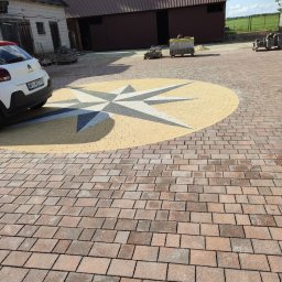 Adam Bąchort - Ułożona kostka brukowa w kształcie rozety w kolorach beżu, szarości i brązu. W tle budynki gospodarcze i zaparkowany samochód. Widok z perspektywy.