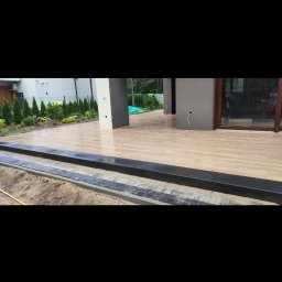 Adam Bąchort - Jasny taras z płytek imitujących drewno, obrzeża z ciemnego kamienia, nowoczesny dom z dużymi oknami i zadbanym ogrodem w tle. Widok z poziomu gruntu.