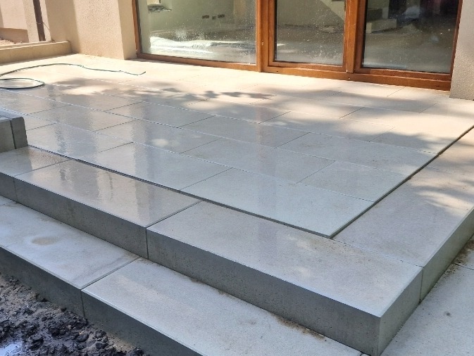 Jasnoszary taras kamienny z dwoma stopniami, wykonany z prostokątnych płyt. Widoczne przesuwne drzwi tarasowe z brązowymi ramami i fragment węża ogrodowego.