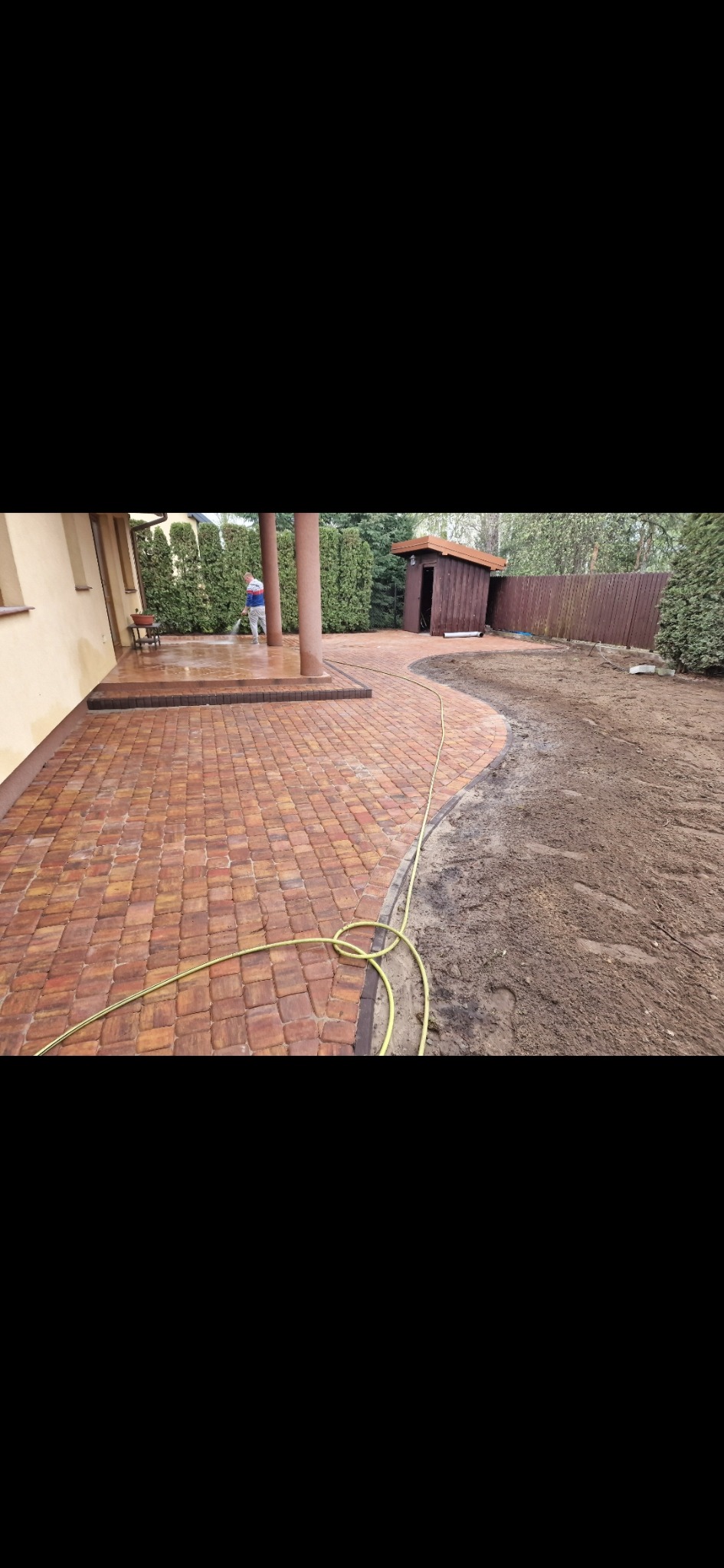 Kaskadowy taras z kostki brukowej w odcieniach brązu, z kolumnami i zielenią w tle. Wąż ogrodowy leży na kostce. Osoba myje kostkę. Widoczny fragment ogrodu.