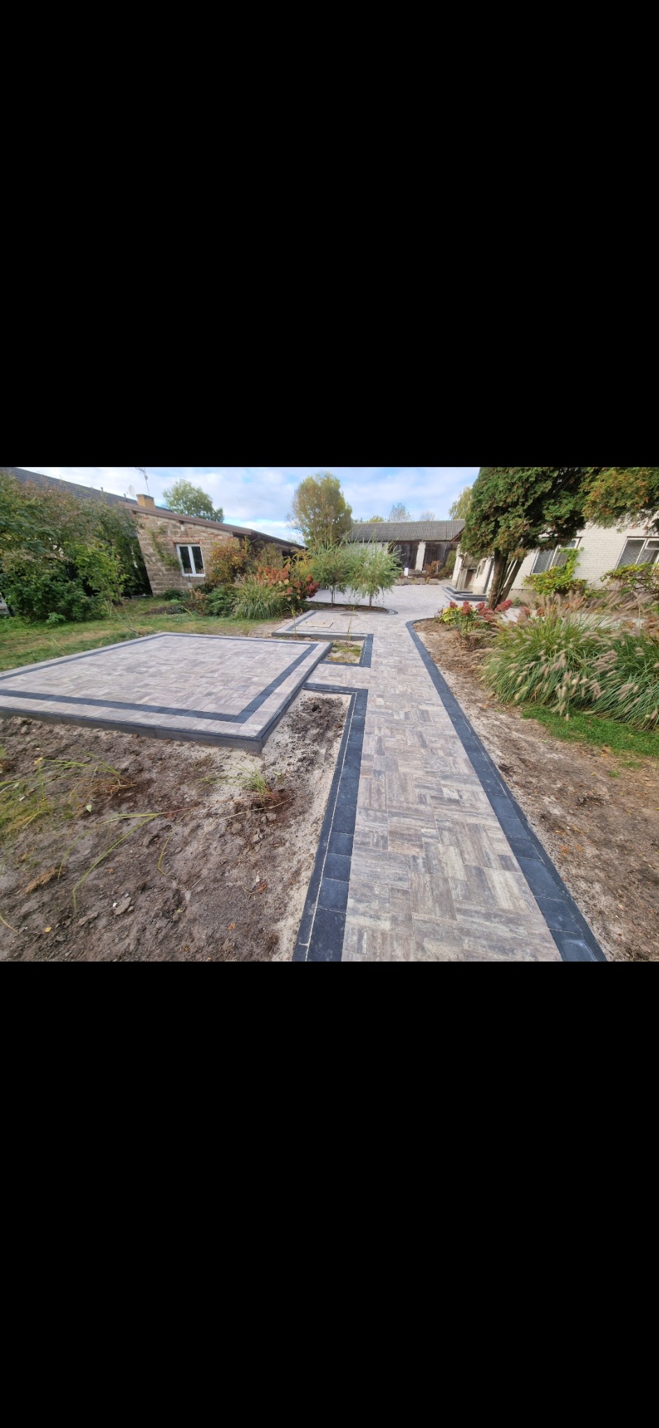 Ułożona kostka brukowa w geometryczny wzór, obramowana ciemniejszymi kostkami, tworząca ścieżkę i plac na posesji z zielenią w tle.