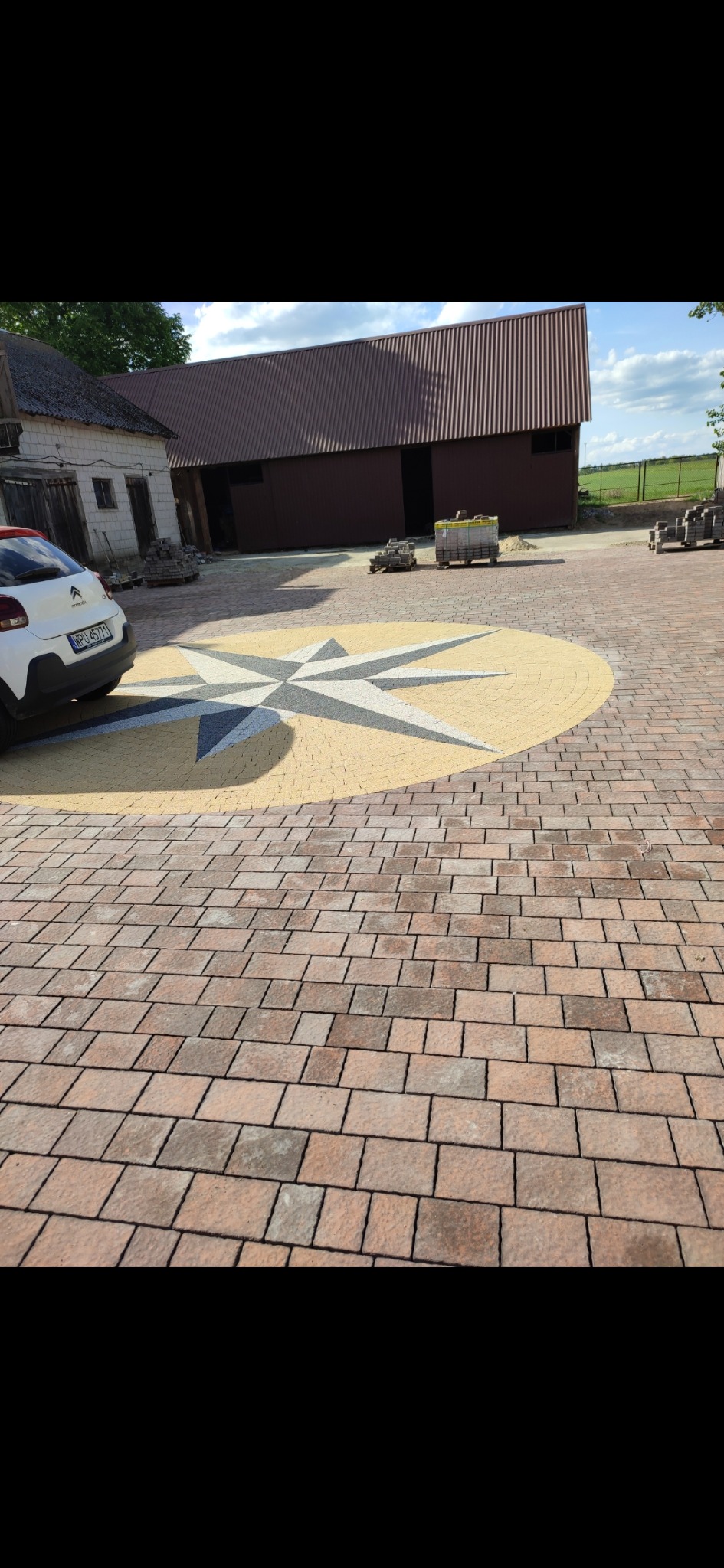 Ułożona kostka brukowa w kształcie rozety w kolorach beżu, szarości i brązu. W tle budynki gospodarcze i zaparkowany samochód. Widok z perspektywy.