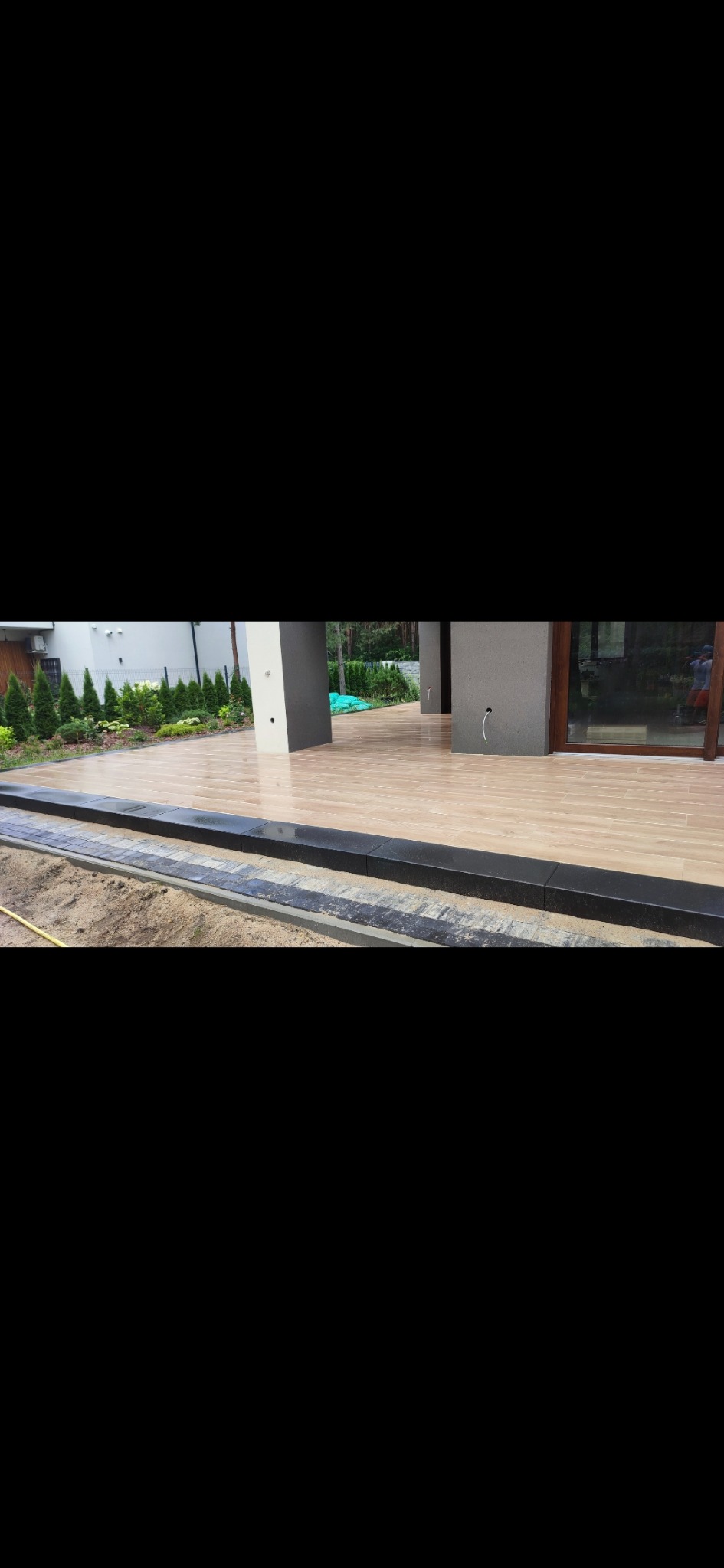 Jasny taras z płytek imitujących drewno, obrzeża z ciemnego kamienia, nowoczesny dom z dużymi oknami i zadbanym ogrodem w tle. Widok z poziomu gruntu.