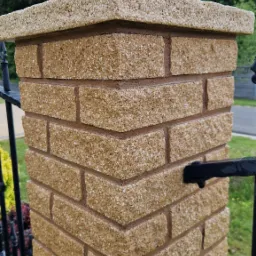 Detal kolumny ogrodzeniowej z beżowej kostki, zwieńczonej jasnym elementem. Widok na fragment ogrodzenia, zieleń i niebo w tle. Precyzyjne wykonanie.