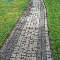 MT Mycie ciśnieniowe - Ścieżka z kostki brukowej, częściowo wyczyszczona, z widocznym sprzętem i odzieżą roboczą na trawniku. Widoczny efekt czyszczenia.