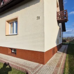 Dwupiętrowy dom z beżową elewacją i numerem 78, z balkonem i kostką brukową. W tle błękitne niebo. Cień osoby fotografującej w lewym dolnym rogu.