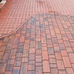 Czerwona kostka brukowa ułożona w dwa wzory: prostokątny i łukowy. W tle zieleń trawnika i fragment czerwonego samochodu.