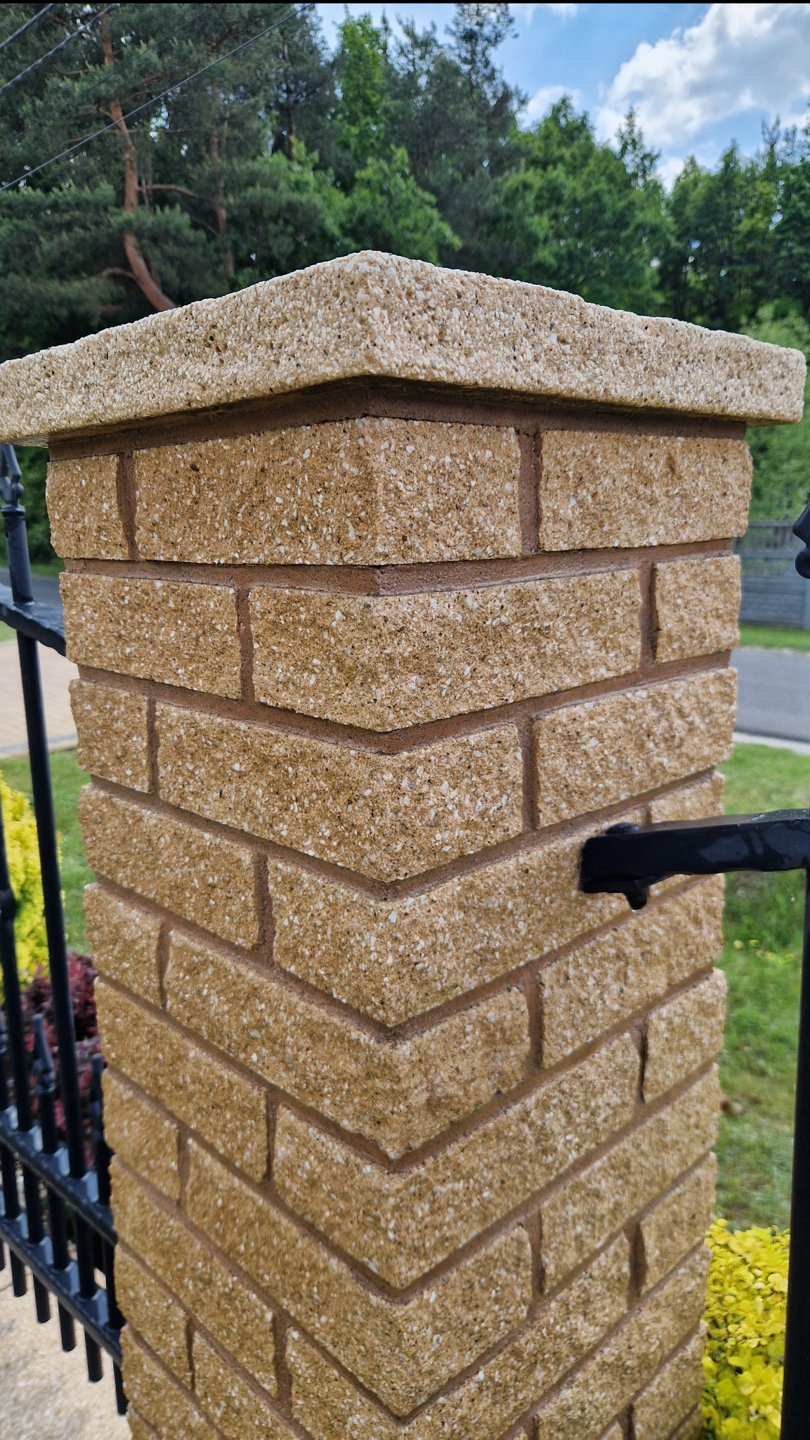 Detal kolumny ogrodzeniowej z beżowej kostki, zwieńczonej jasnym elementem. Widok na fragment ogrodzenia, zieleń i niebo w tle. Precyzyjne wykonanie.