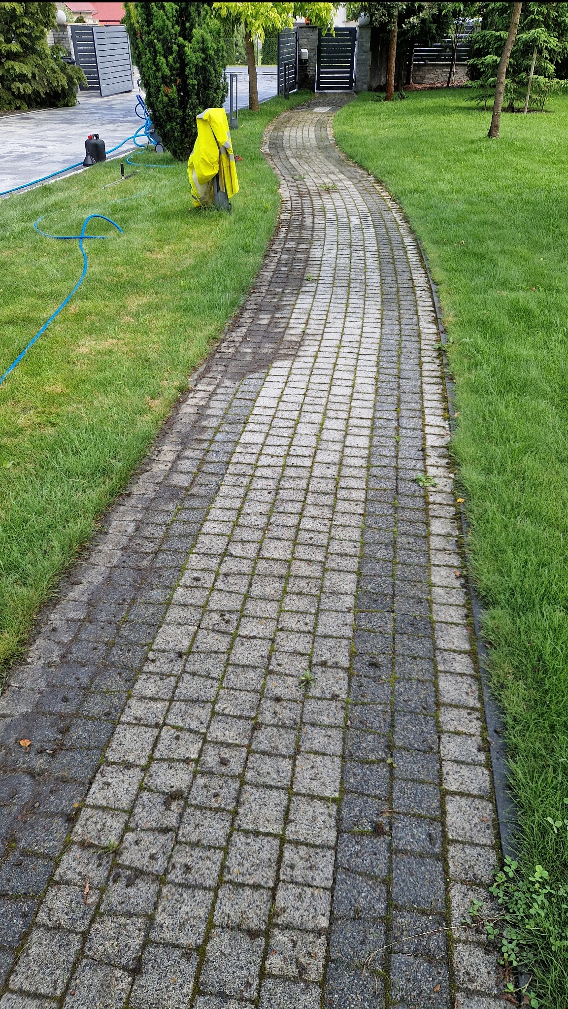 Ścieżka z kostki brukowej, częściowo wyczyszczona, z widocznym sprzętem i odzieżą roboczą na trawniku. Widoczny efekt czyszczenia.
