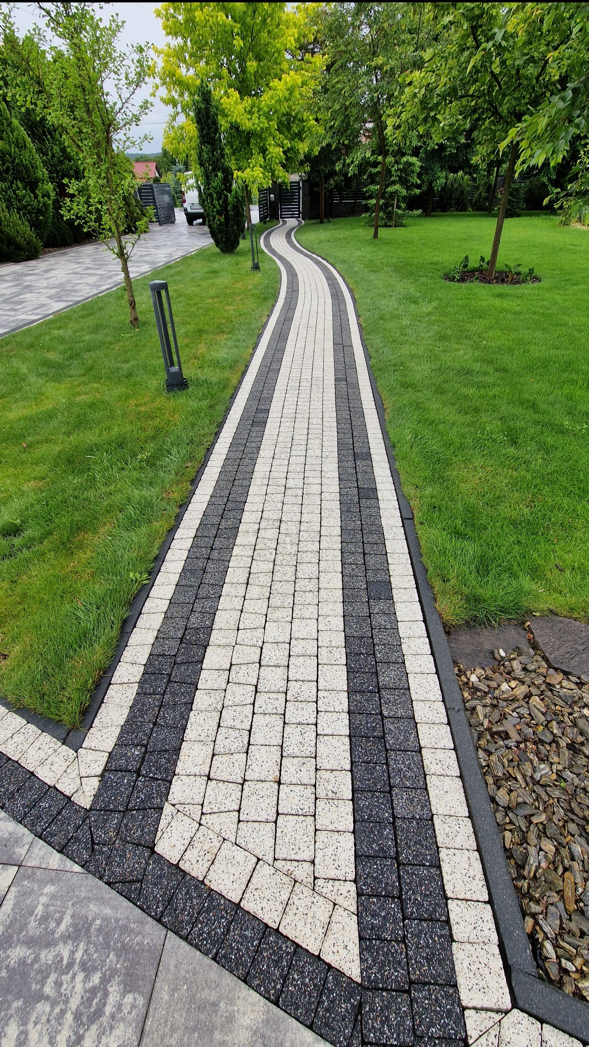 Kręta ścieżka z kostki brukowej w odcieniach szarości i bieli, wijąca się przez trawnik, z lampami po bokach i drzewami w tle. Estetyczne wykończenie ogrodu.