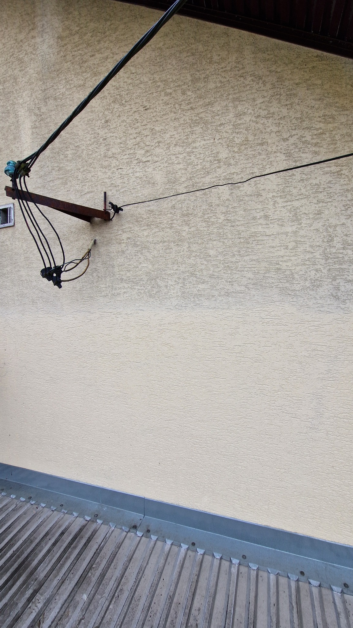 Fragment elewacji budynku z widocznym zabrudzeniem na ścianie, na której zamontowane są kable i metalowe elementy. U dołu widoczny fragment dachu.