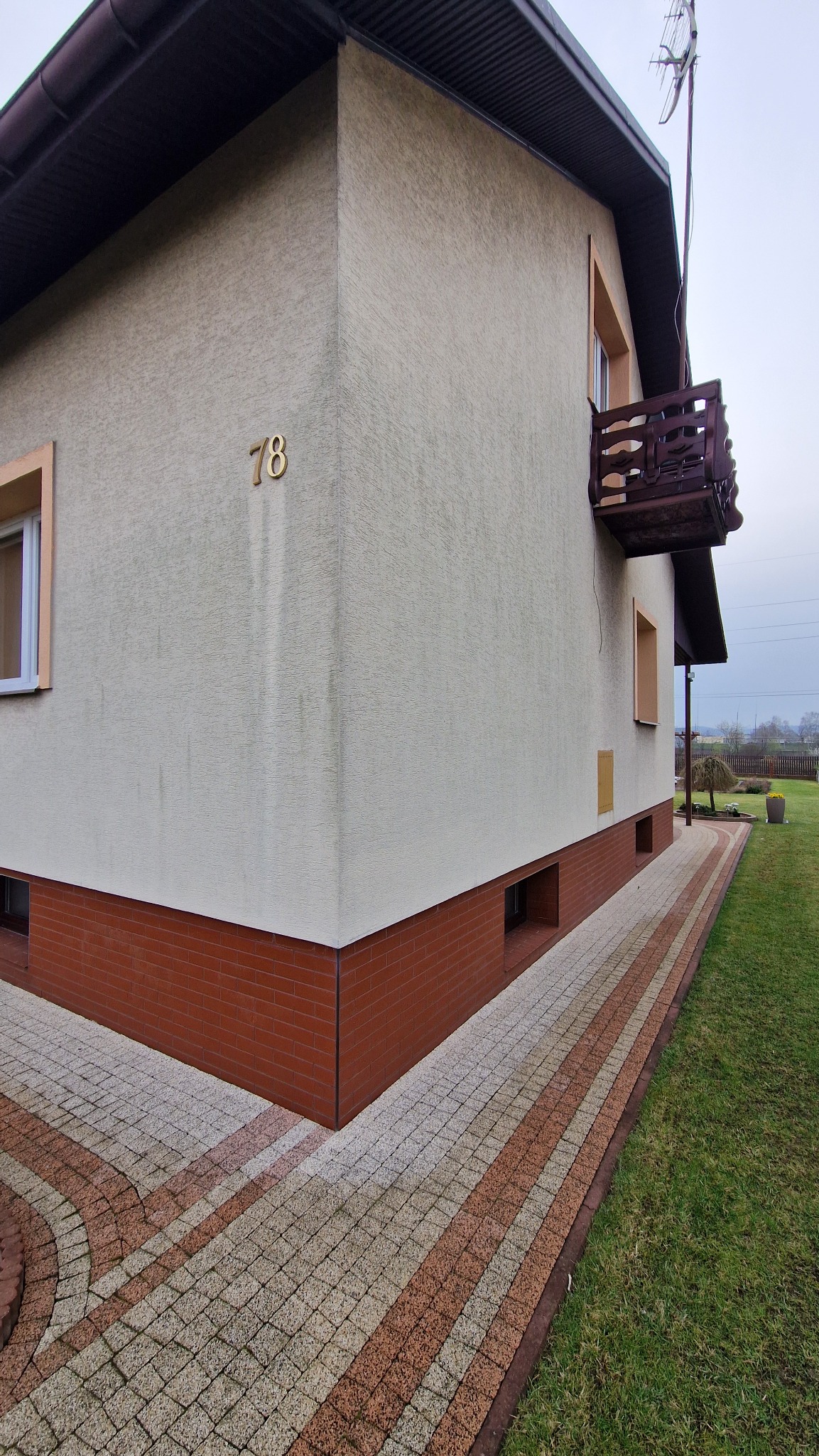 Częściowy widok elewacji domu z beżowym tynkiem, numerem 78, balkonem z brązową balustradą i kostką brukową w odcieniach brązu i szarości.
