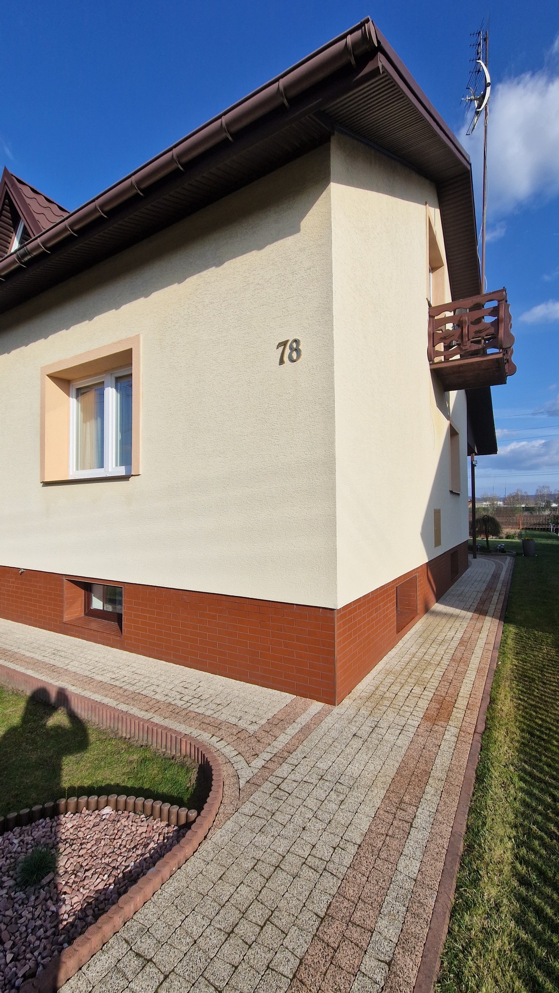 Dwupiętrowy dom z beżową elewacją i numerem 78, z balkonem i kostką brukową. W tle błękitne niebo. Cień osoby fotografującej w lewym dolnym rogu.