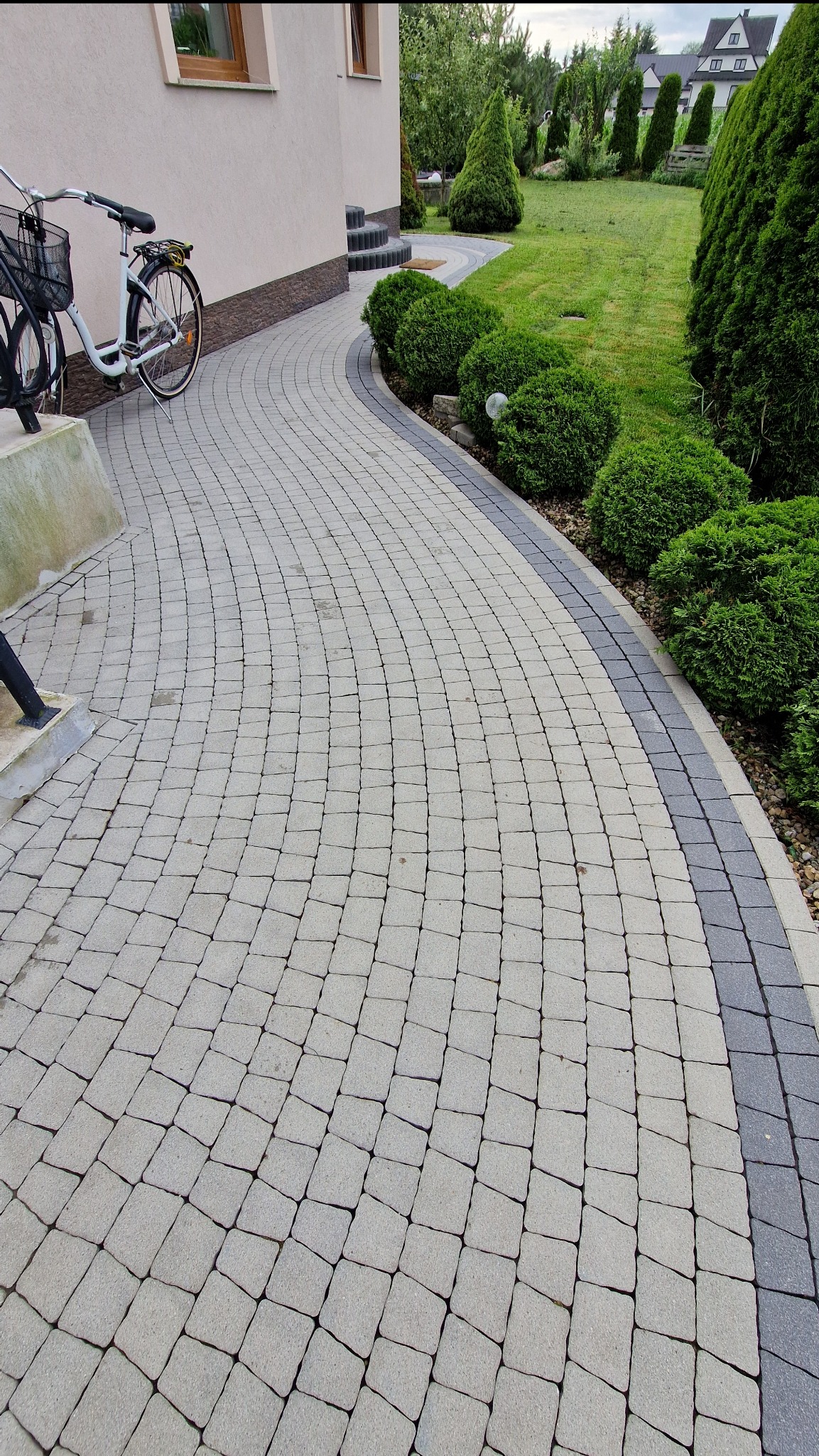 Zakrzywiony chodnik z kostki brukowej w odcieniach szarości, obok domu z rowerem i zadbanym trawnikiem z krzewami. Widok z perspektywy.