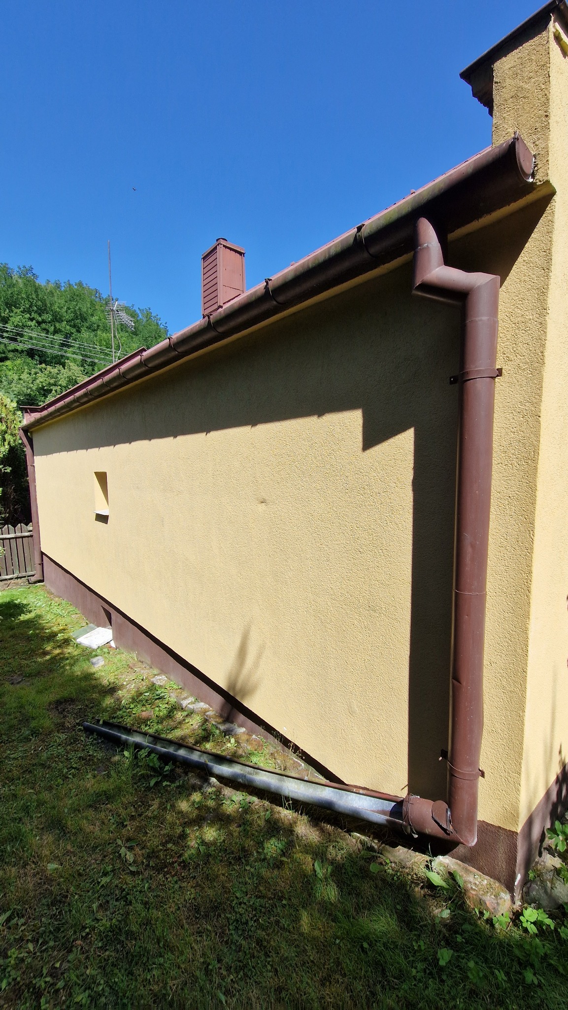 Żółta elewacja budynku z brązowymi rynnami w słoneczny dzień. Widoczny fragment trawnika i niebieskie niebo. Rynna odchodzi od rogu budynku.