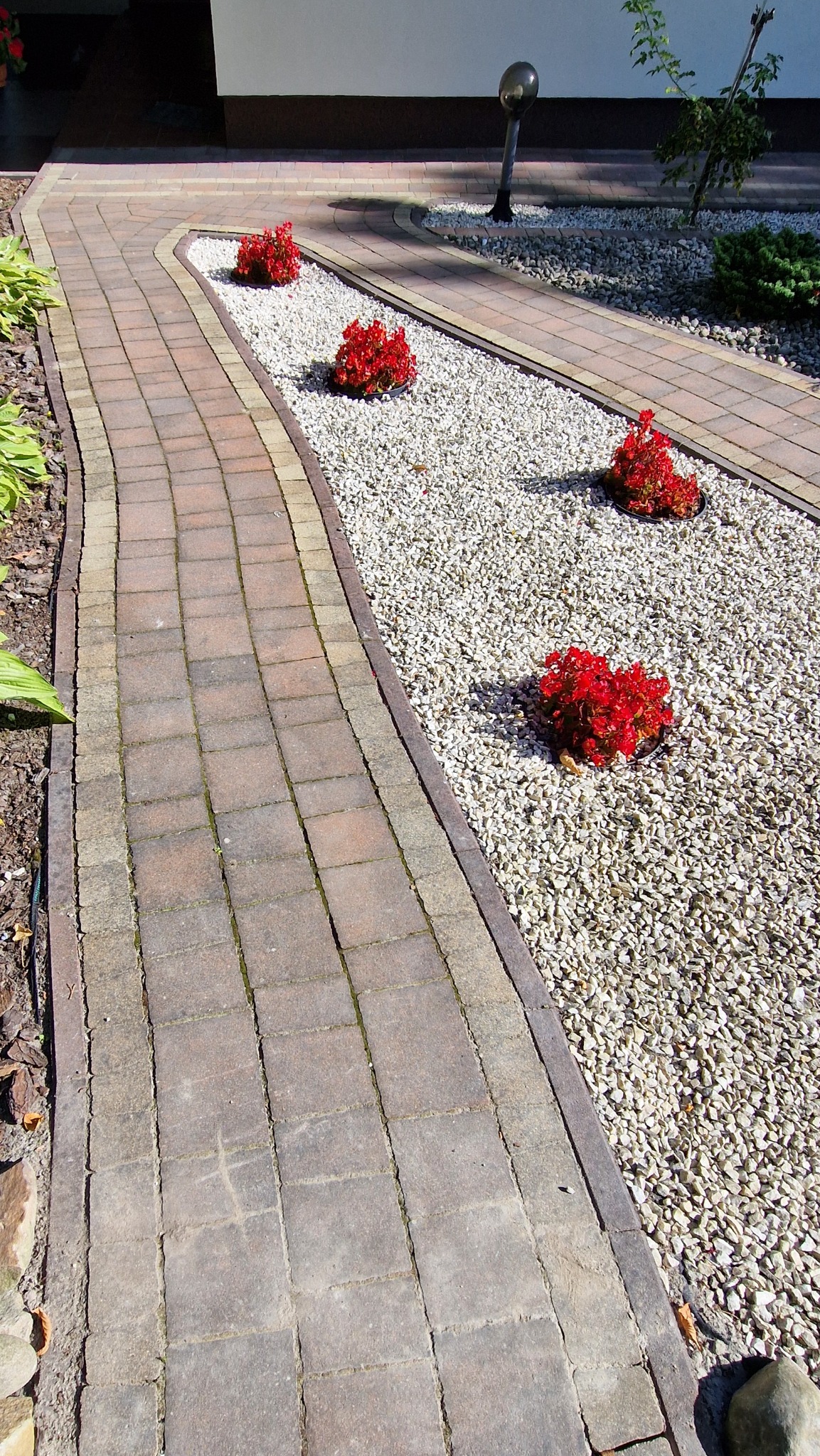 Ścieżka z kostki brukowej, obrzeża z kamienia, rabaty z białym grysem i czerwonymi kwiatami. Oświetlenie ogrodowe. Ujęcie z góry.