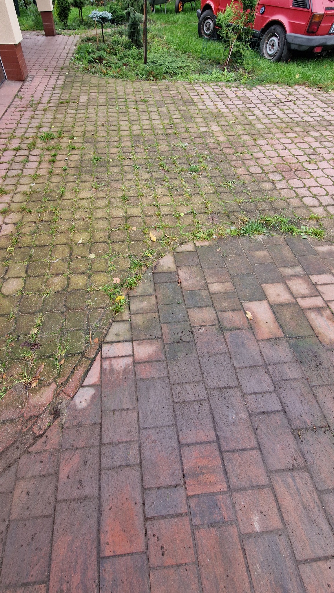 Częściowo wyczyszczona kostka brukowa na zewnątrz budynku, z widoczną różnicą w kolorze i czystości między umytą a zabrudzoną powierzchnią. W tle zieleń i czerwony samochód.