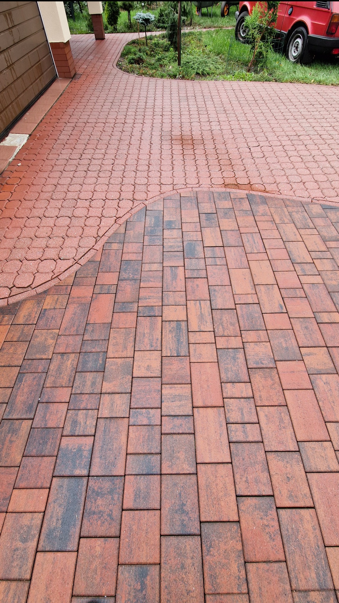 Czerwona kostka brukowa ułożona w dwa wzory: prostokątny i łukowy. W tle zieleń trawnika i fragment czerwonego samochodu.