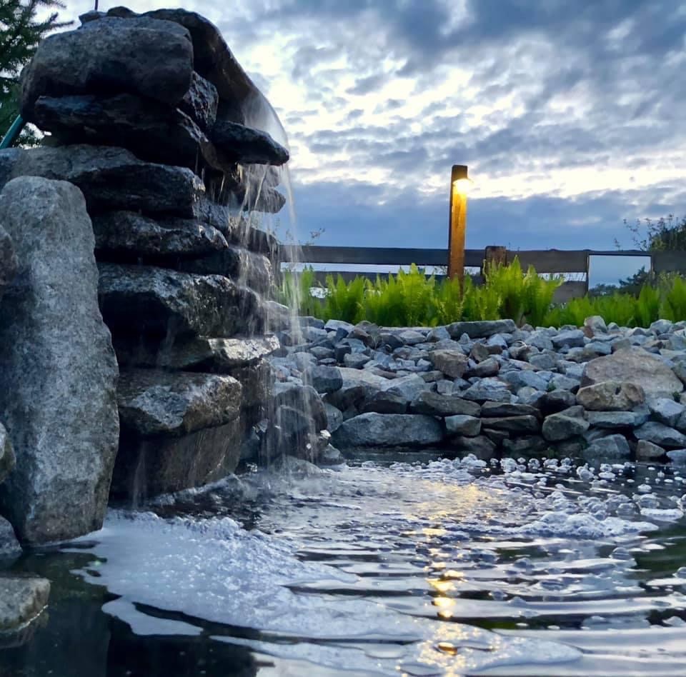 Kaskada wodna z szarego kamienia w ogrodzie, woda spływa do oczka wodnego, w tle drewniane ogrodzenie i oświetlenie ogrodowe pod wieczornym niebem.