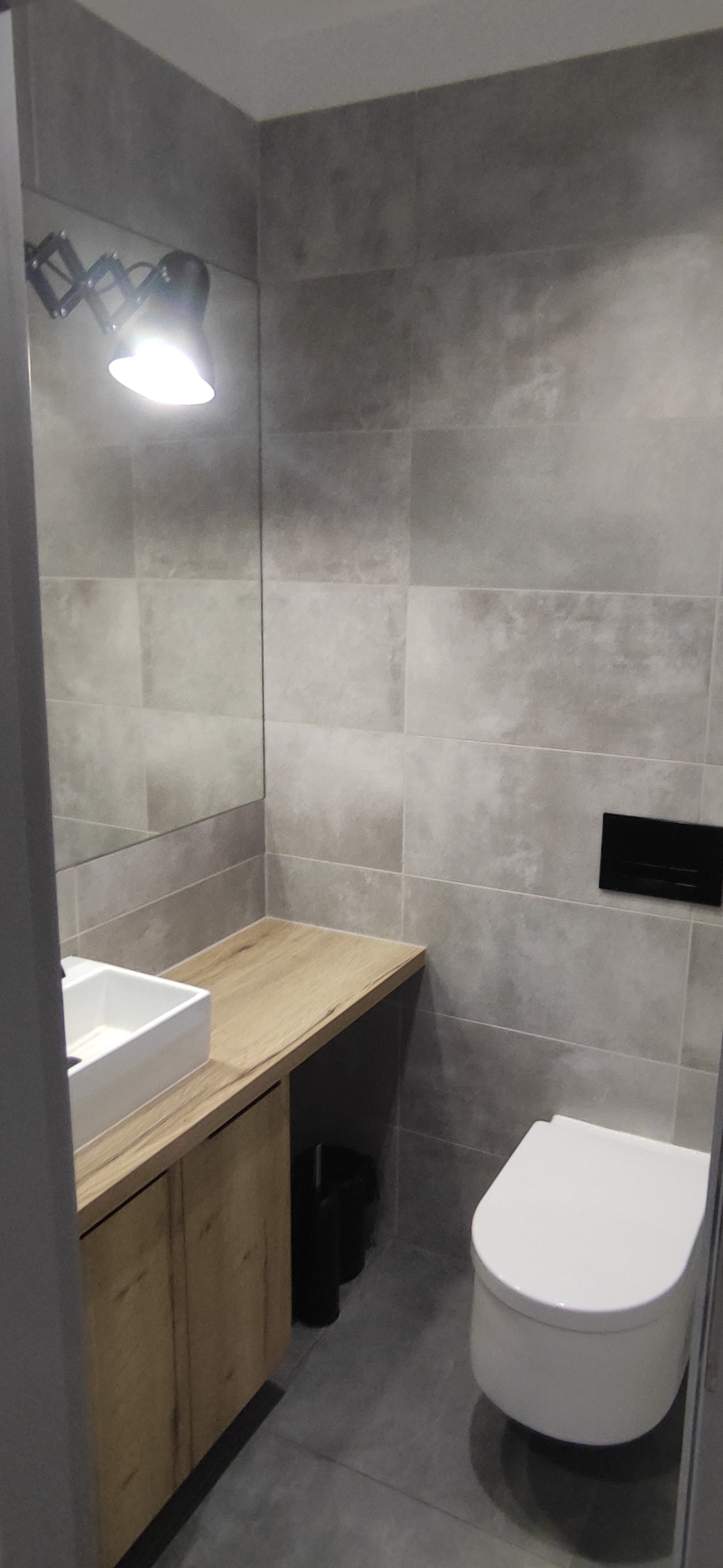 Nowoczesna łazienka z szarymi płytkami, podwieszaną toaletą, szafką pod umywalką z drewnianym blatem i regulowaną lampą nad lustrem.