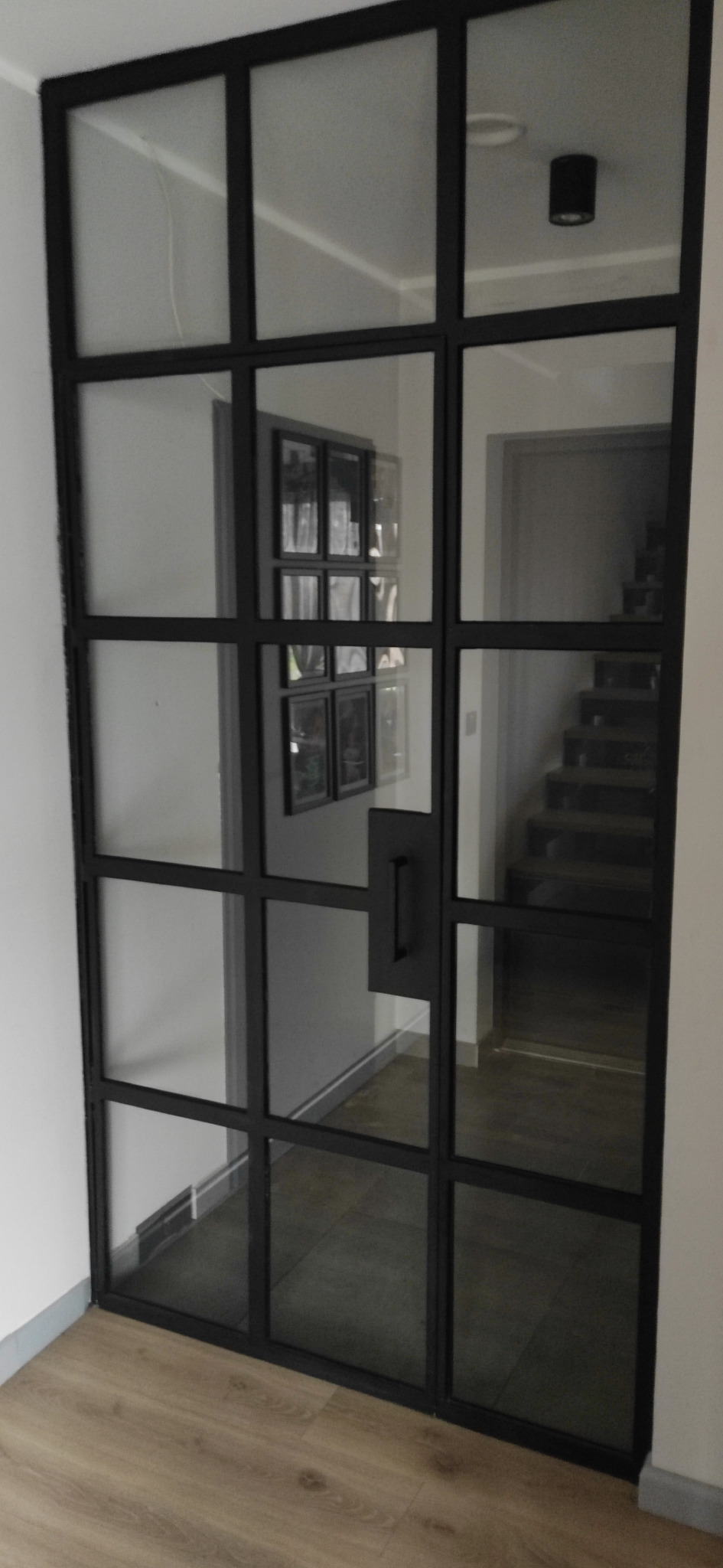 Szklane drzwi loftowe z czarną ramą, widok na korytarz z schodami i galerią zdjęć na ścianie.