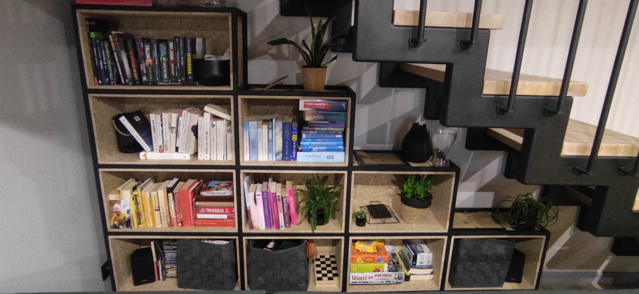 Regał modułowy z płyty OSB z czarnymi obramowaniami, wypełniony książkami, roślinami doniczkowymi i grami planszowymi, ustawiony pod schodami z drewnianymi stopniami i metalową konstrukcją.