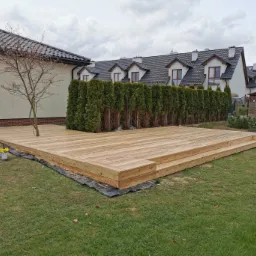 Nowo wybudowany taras z jasnego drewna otoczony trawnikiem, z widocznym młodym drzewkiem wyrastającym z tarasu, w tle rząd wysokich tuj i domy jednorodzinne pod pochmurnym niebem.
