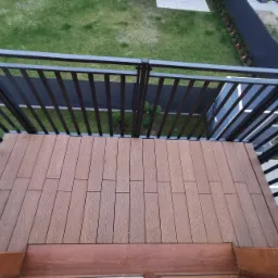 Nowoczesny balkon z brązową podłogą z desek kompozytowych i czarną metalową balustradą, widok z góry na zielony trawnik i ścieżkę.