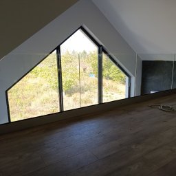 MS-PRO Marek Szczepanek - Szklana balustrada na antresoli pod skosem dachu, widok na okno trójkątne z czarną ramą i zielony krajobraz za nim. Na podłodze leży zwinięta lina.