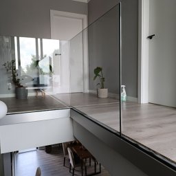 MS-PRO Marek Szczepanek - Szklana balustrada w Łodzi, z widokiem na dół, gdzie widać stół i krzesła. Na górnym poziomie, za balustradą, znajdują się rośliny doniczkowe i białe drzwi.