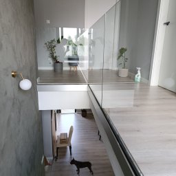 MS-PRO Marek Szczepanek - Nowoczesna balustrada szklana z widokiem na schody, dwa psy na schodach, minimalistyczne wnętrze z roślinami doniczkowymi i lampą ścienną w kolorze złota.