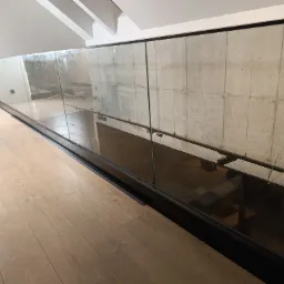 Szklana balustrada z czarnymi elementami mocującymi i poręczą, zamontowana na antresoli z betonową ścianą w tle, widok z parkietu.