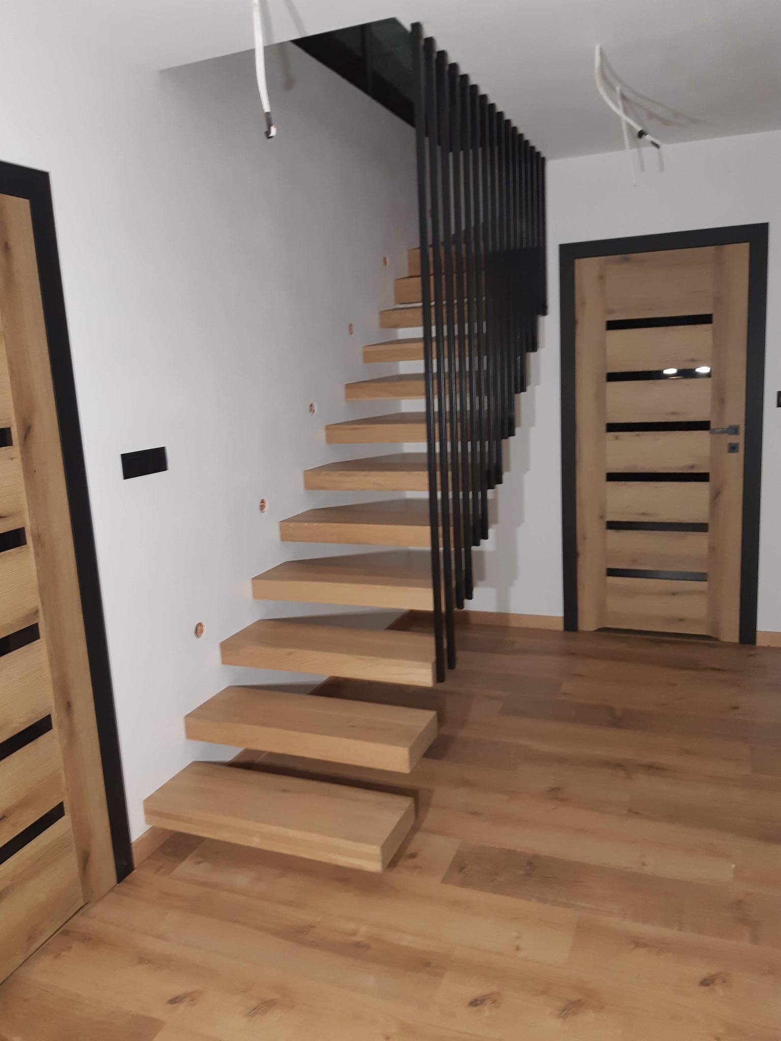Drewniane schody wspornikowe z czarną, pionową balustradą, zamontowane w nowoczesnym wnętrzu z drewnianą podłogą i drzwiami z czarnymi akcentami.