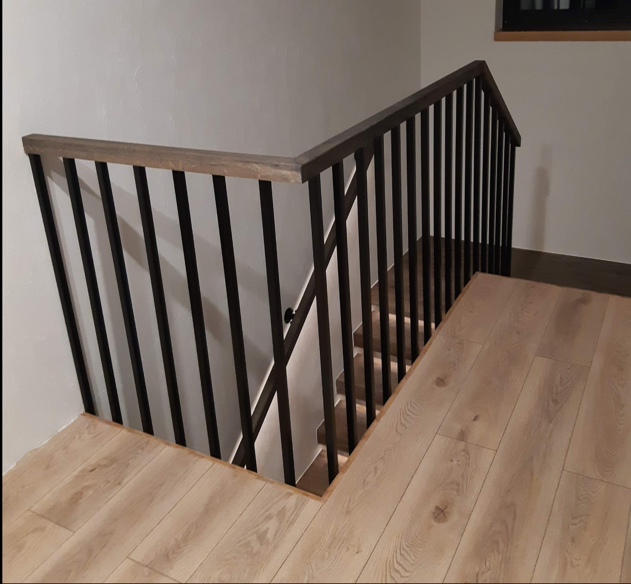 Nowoczesne schody z drewnianymi stopniami i czarną, metalową balustradą z drewnianym poręczą, widok z góry na jasną podłogę.