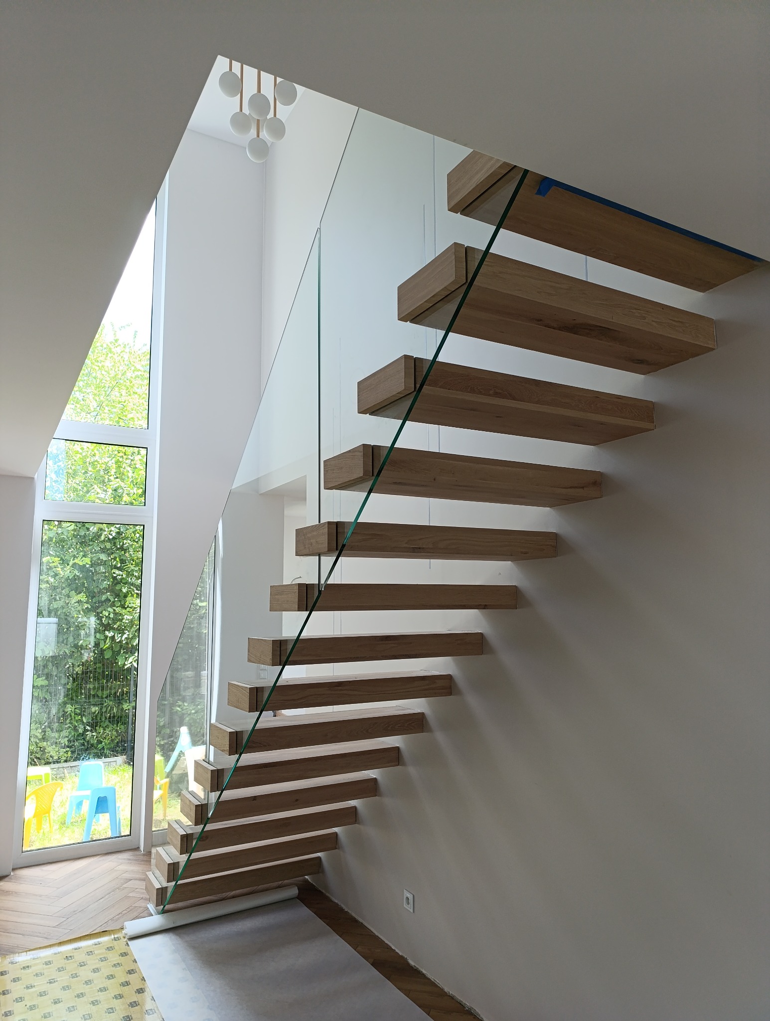 Nowoczesne schody z drewnianymi stopniami i szklaną balustradą w jasnym wnętrzu z dużym oknem.