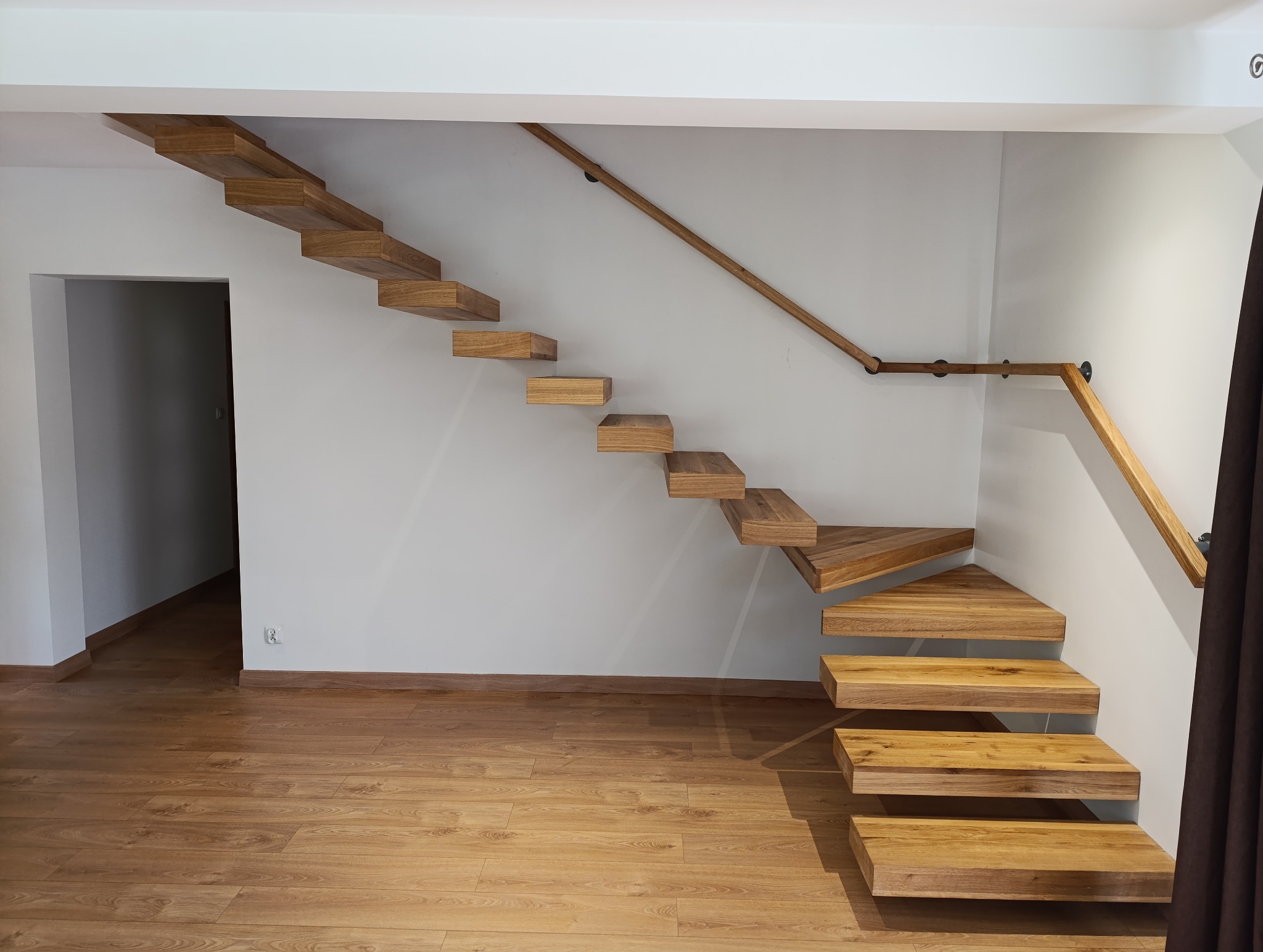 Nowoczesne, drewniane schody wspornikowe z drewnianą poręczą zamocowaną do ściany, widoczne z dołu, na tle białej ściany i drewnianej podłogi.
