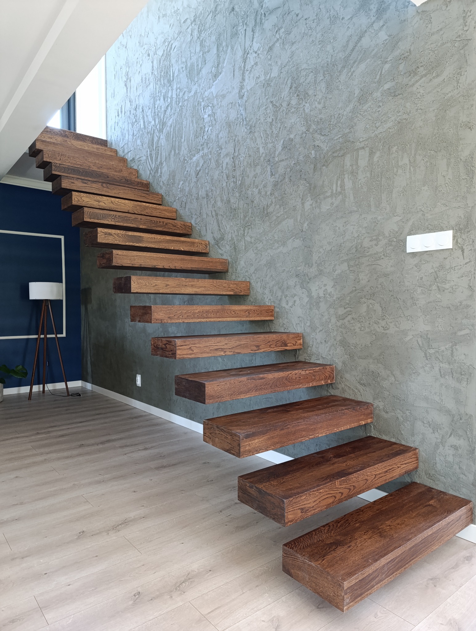 Drewniane schody wspornikowe o ciemnym wybarwieniu, zamontowane przy szarej ścianie z fakturą betonu, w tle widoczna lampa na trójnogu i granatowa ściana.