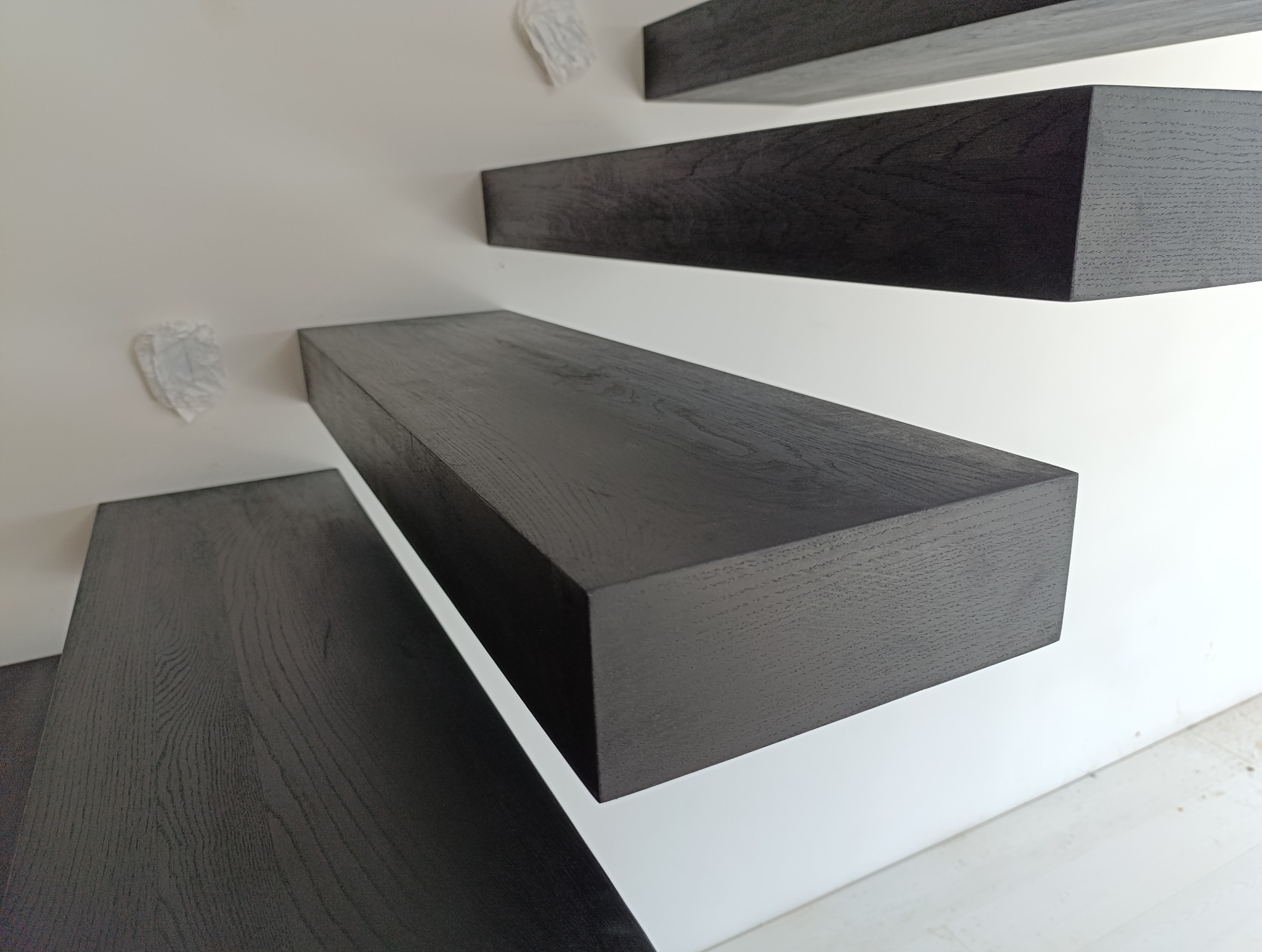 Minimalistyczne, wiszące stopnie schodów metalowych o ciemnym, drewnopodobnym wykończeniu, zamocowane na białej ścianie.