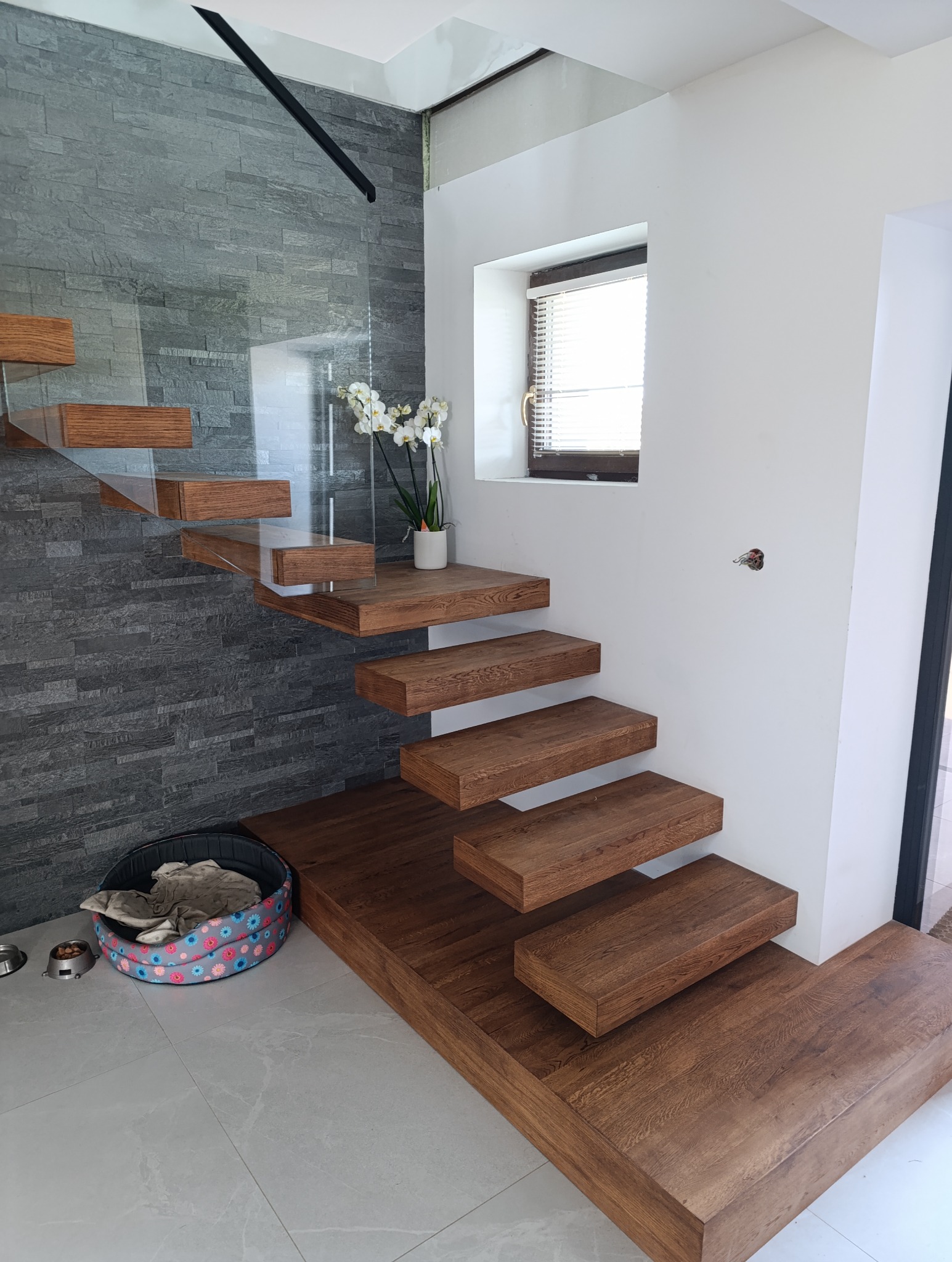 Nowoczesne, drewniane schody wspornikowe z szklaną balustradą, oświetlone naturalnym światłem z okna, z widoczną dekoracją kwiatową i legowiskiem dla zwierząt u dołu schodów.