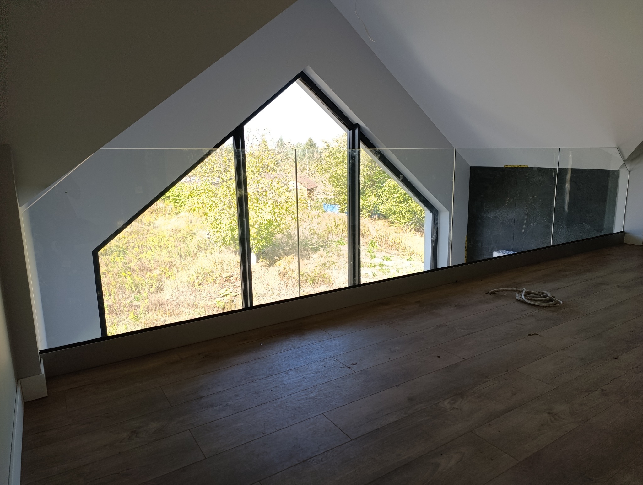 Szklana balustrada na antresoli pod skosem dachu, widok na okno trójkątne z czarną ramą i zielony krajobraz za nim. Na podłodze leży zwinięta lina.
