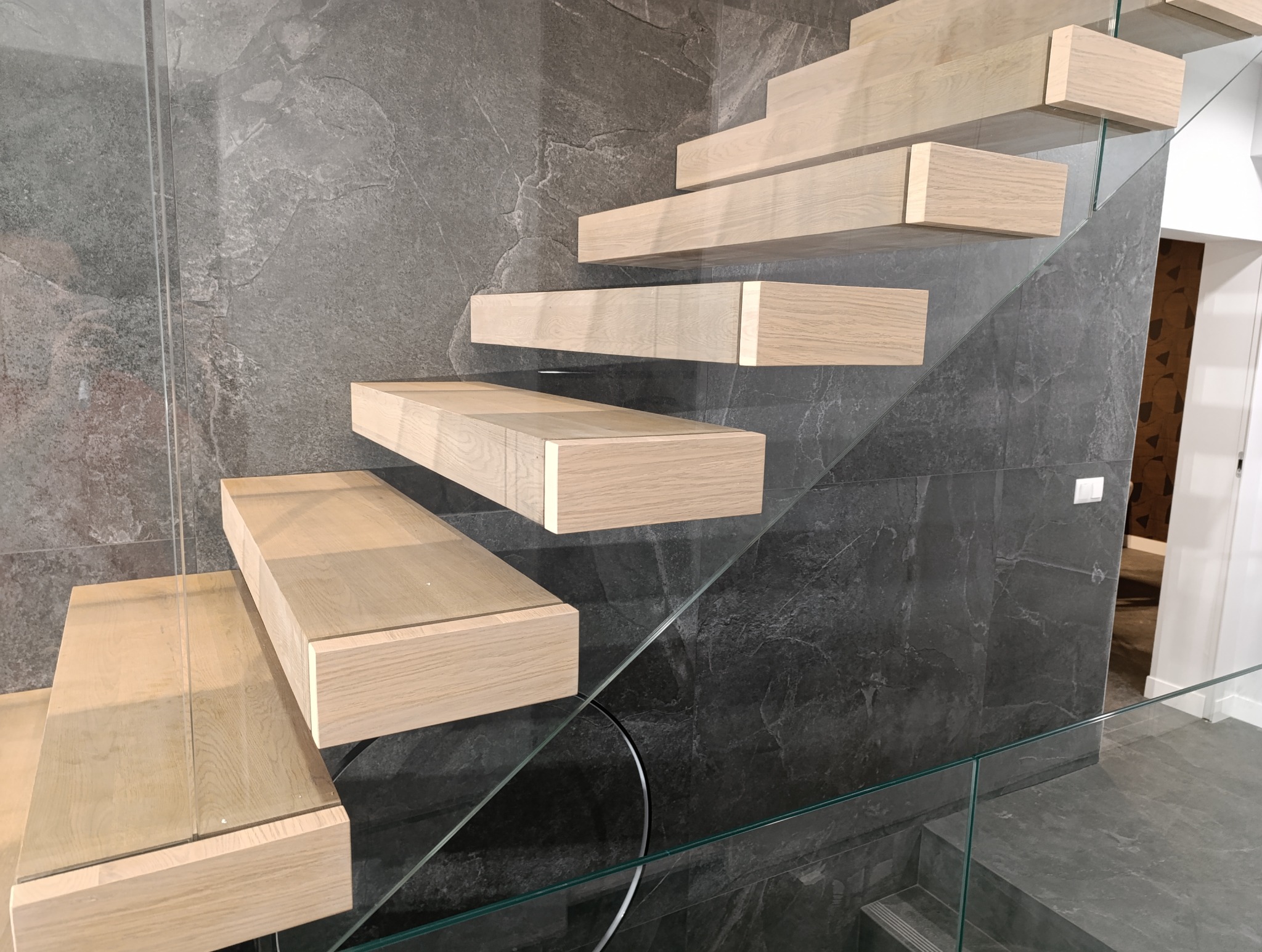 Nowoczesne schody wspornikowe z jasnego drewna, zamocowane do ściany z szarego kamienia, z balustradą ze szkła hartowanego.