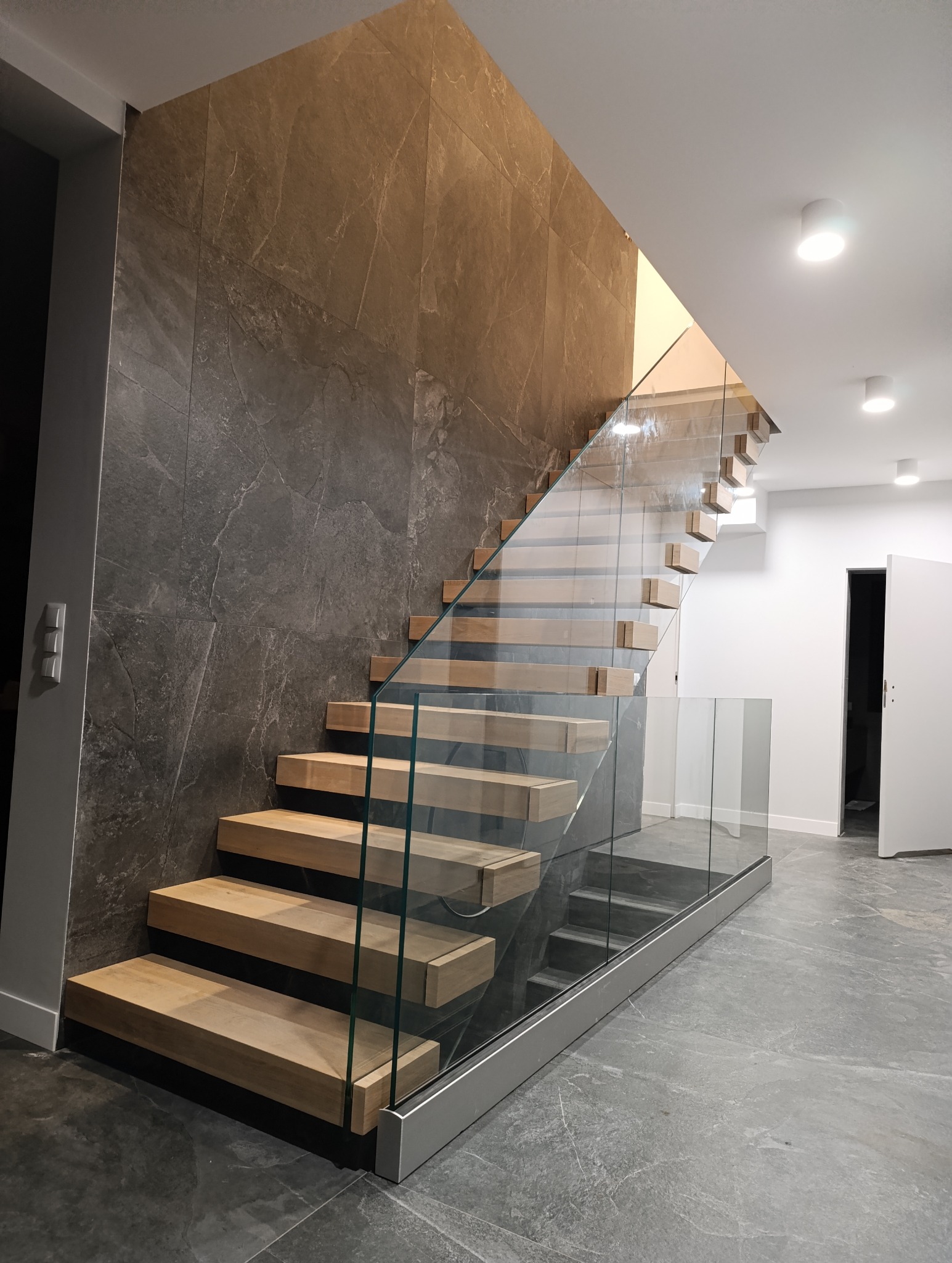 Nowoczesne schody z drewnianymi stopniami i szklaną balustradą, widok z dołu, szara ściana z efektem kamienia w tle.