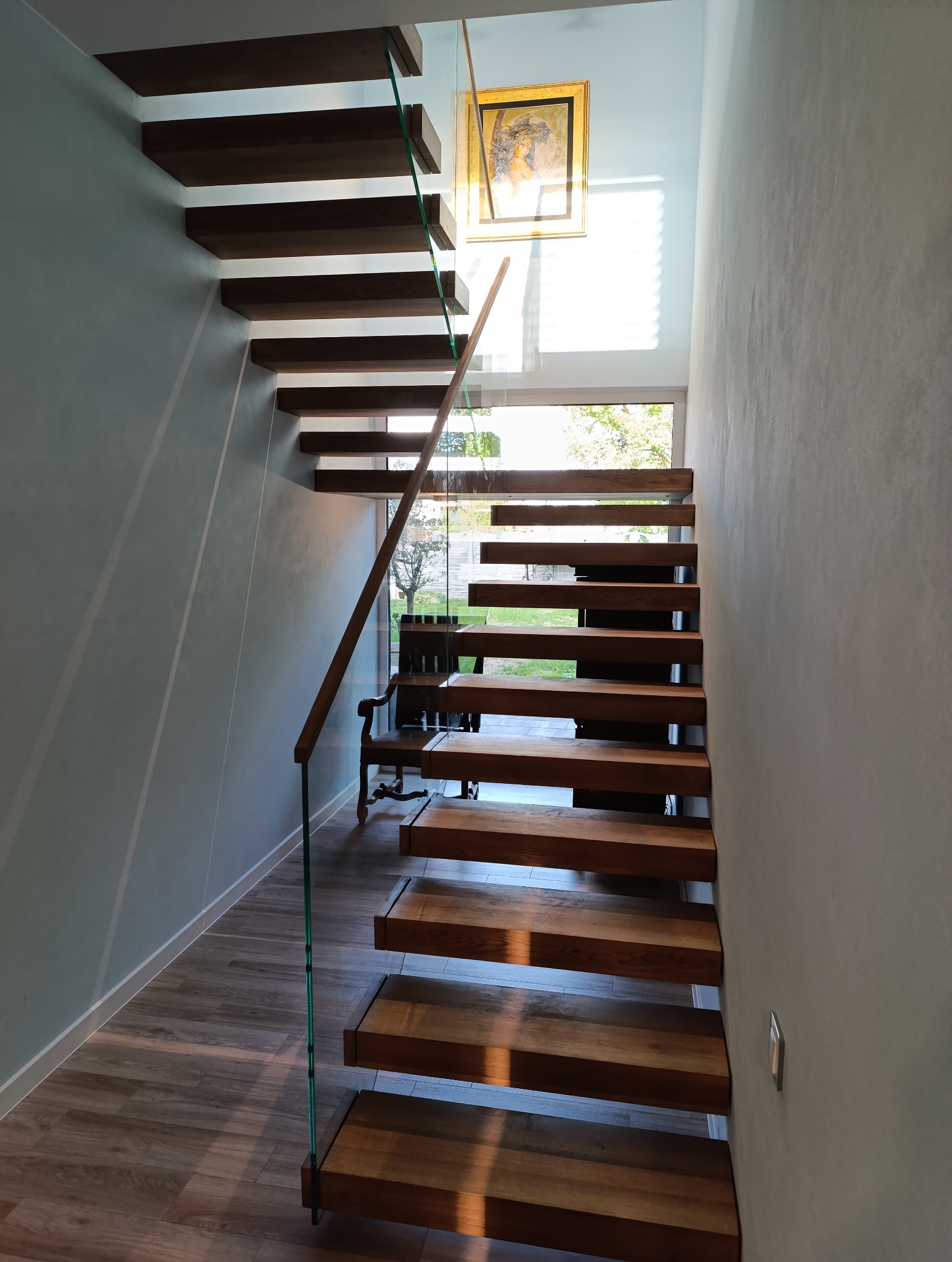 Nowoczesne schody drewniane z balustradą ze szkła, stopnie wspornikowe, widok z dołu ku górze, w tle obraz w złotej ramie i okno z widokiem na zieleń.