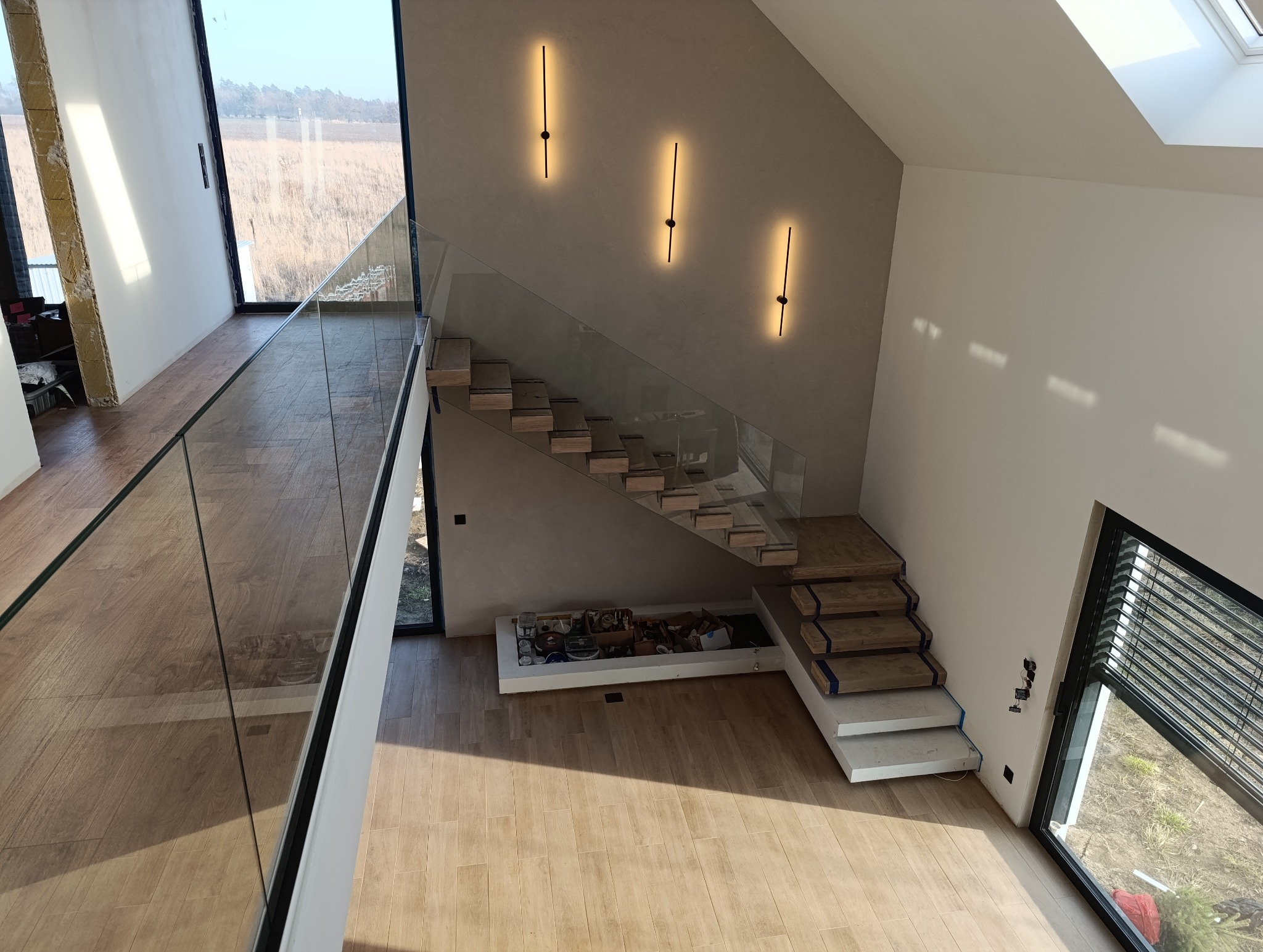 Wnętrze domu z drewnianymi schodami wspornikowymi zabezpieczonymi taśmą, szklaną balustradą na antresoli, minimalistycznymi lampami ściennymi i dużymi oknami.