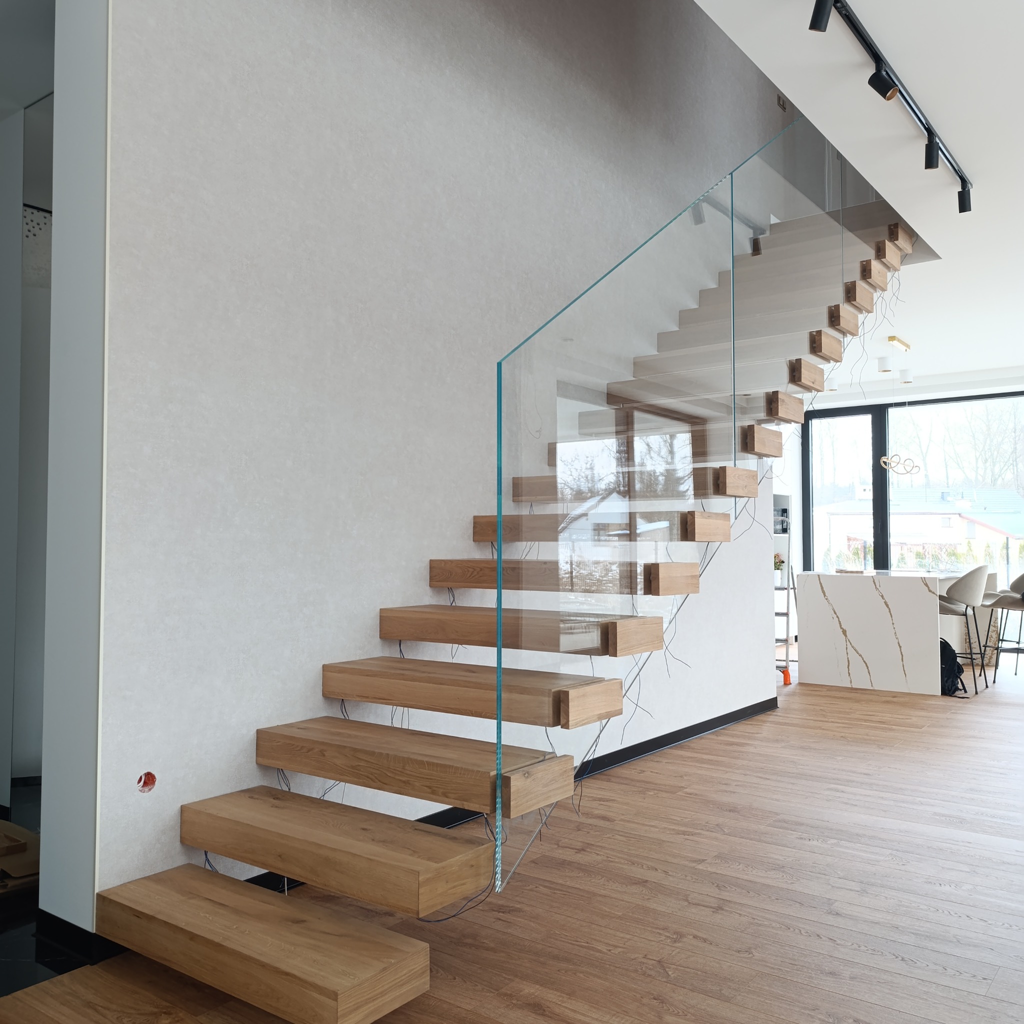 Nowoczesne schody z drewnianymi stopniami i szklaną balustradą, widoczne przewody elektryczne pod stopniami, oświetlenie szynowe na suficie, w tle minimalistyczna kuchnia.