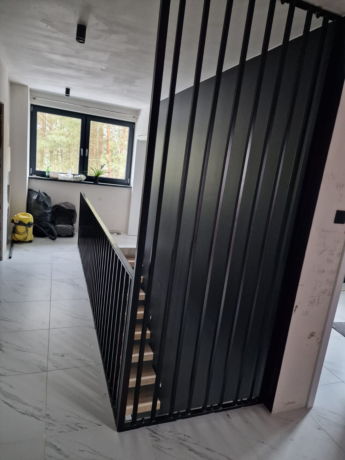 Nowoczesne schody z czarną metalową balustradą z pionowych prętów, widok z dołu na drewniane stopnie, w tle okno z widokiem na las i sprzęt budowlany.