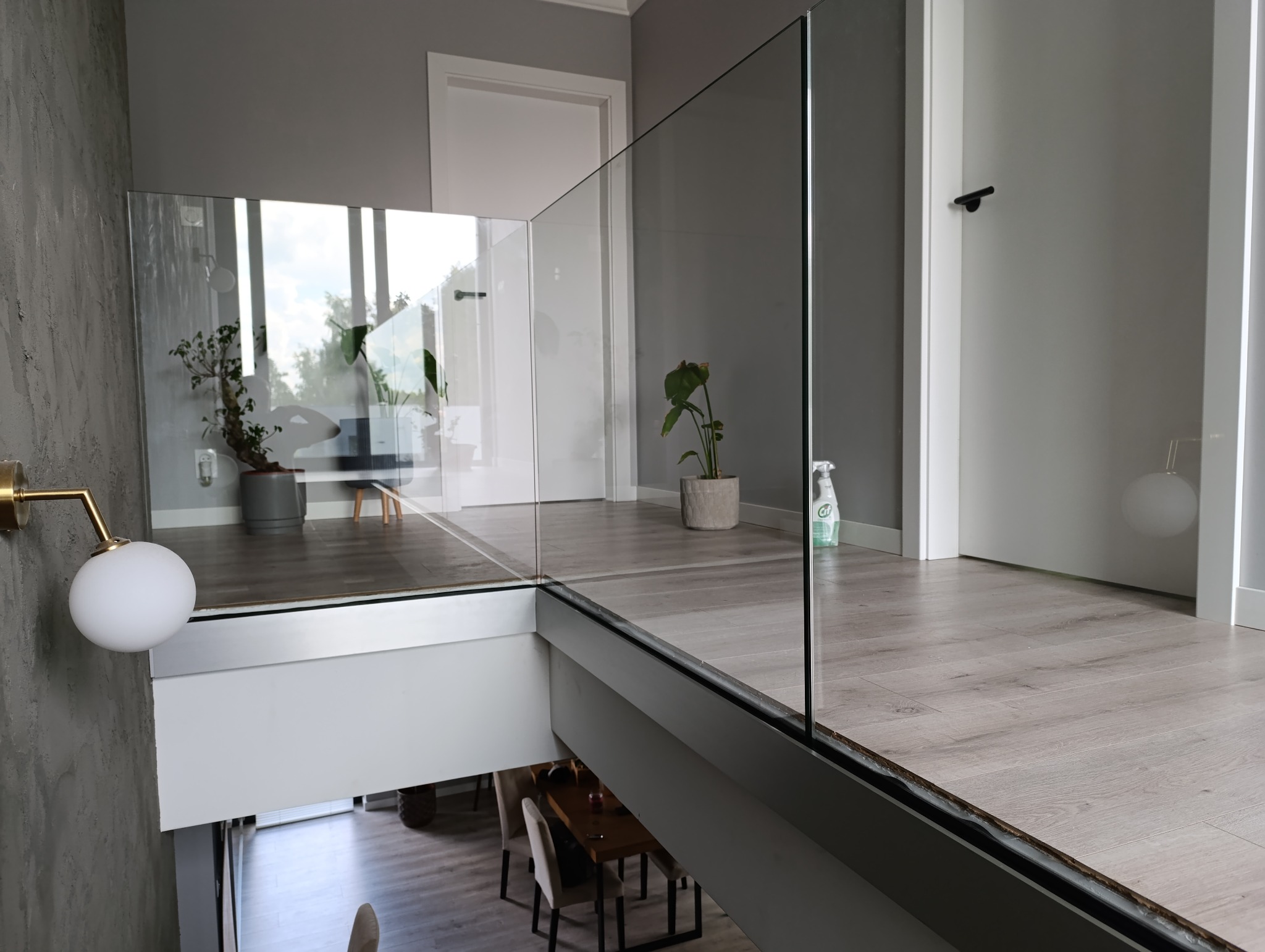 Szklana balustrada w Łodzi, z widokiem na dół, gdzie widać stół i krzesła. Na górnym poziomie, za balustradą, znajdują się rośliny doniczkowe i białe drzwi.