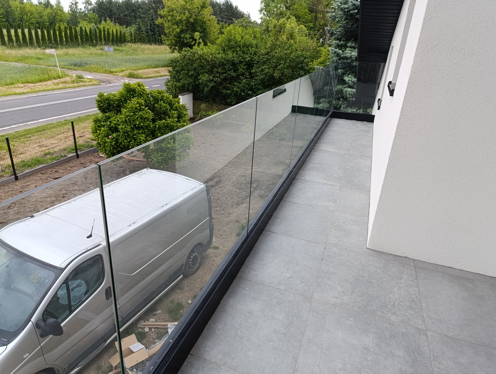 Nowoczesna balustrada szklana na balkonie z widokiem na ulicę i zaparkowany samochód dostawczy.