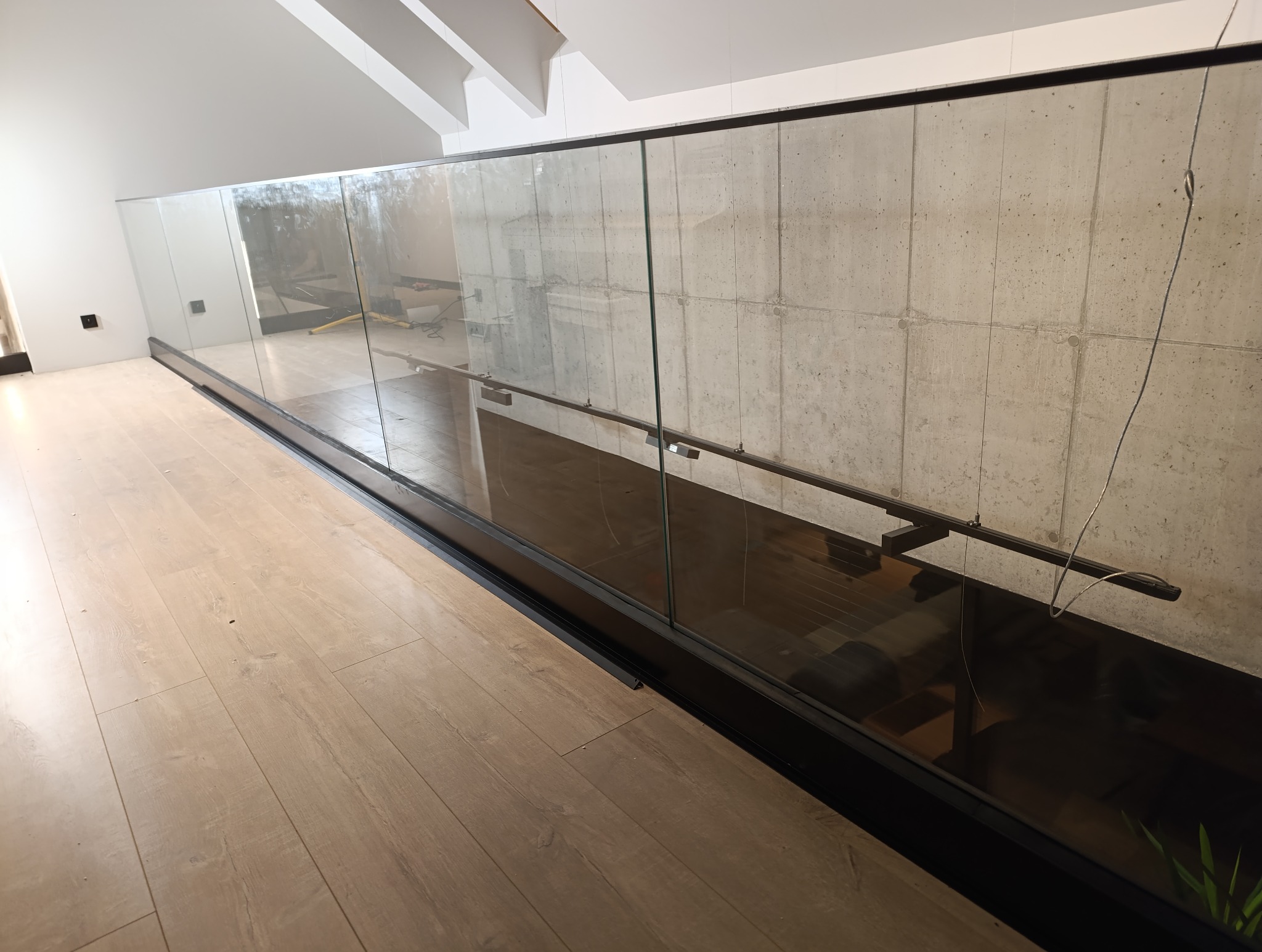 Szklana balustrada z czarnymi elementami mocującymi i poręczą, zamontowana na antresoli z betonową ścianą w tle, widok z parkietu.